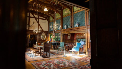 Looking out of the Billiard Room door back into the Great Parlour at Wightwick Manor. The large, wooden table is in the centre of the room on top of red and blue Persian rugs. To the side is the inglenook fireplace with chairs upholstered in William Morris fabrics.